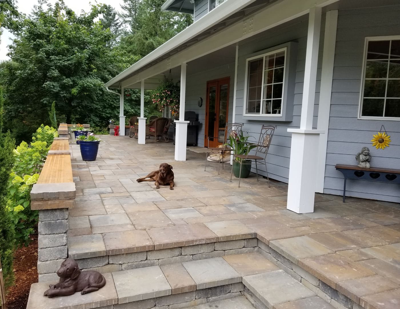 Tan paver porch with white columns, wood-topped seating along a low block wall, potted plants, and a brown dog resting near the door in Brush Prairie