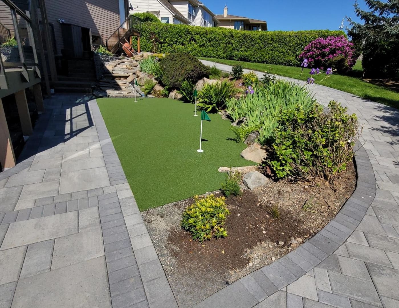 Backyard putting green with three flags, bordered by gray paver paths, boulders, flowering plants, and stone steps in Camas