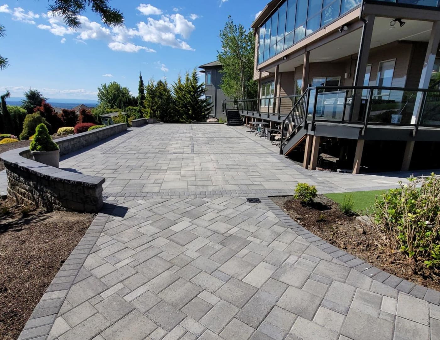Large gray paver patio below a glass-railed deck, with a curved low block wall and dark paver borders in Camas
