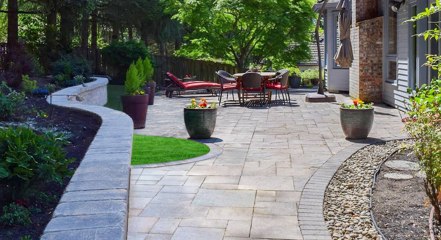 Curved paver walkway and stone retaining wall winding past a sunny paver patio dining area in a Pacific Northwest backyard