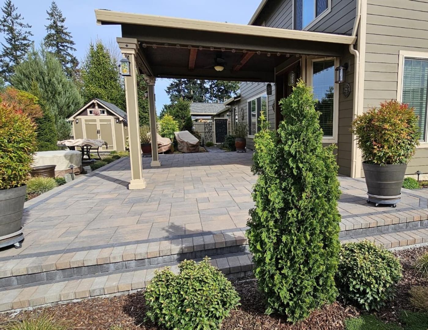 Paver patio beneath a solid-roof cover with a ceiling fan, broad steps, large planters, and evergreen shrubs in Vancouver