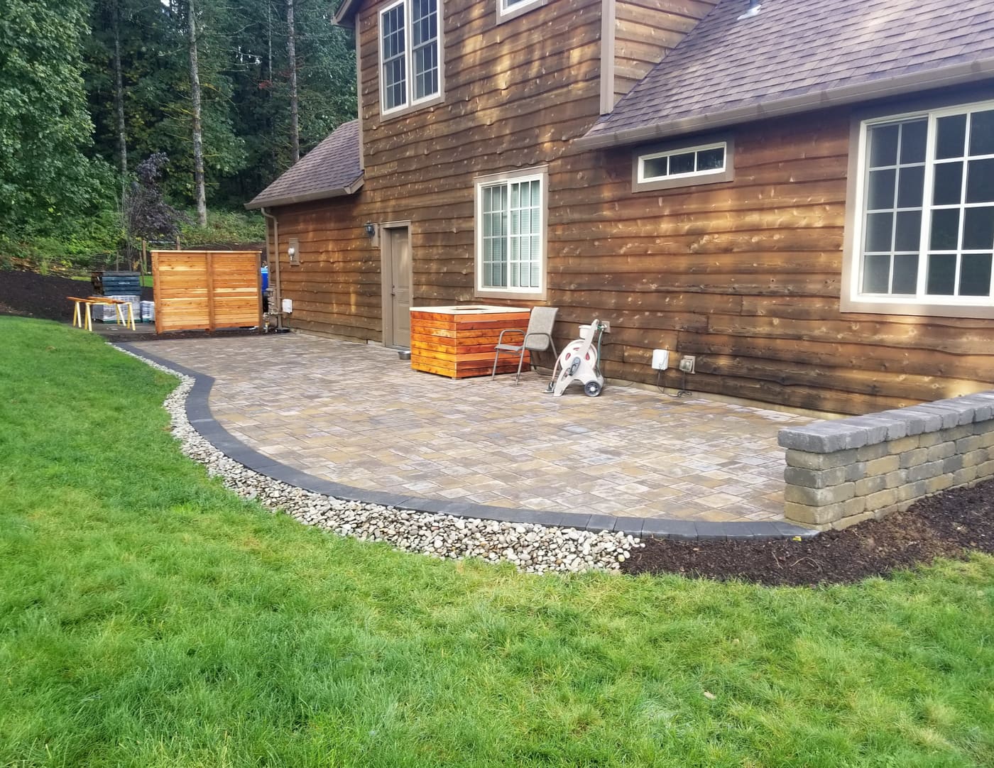 Curved paver patio beside a wood-sided house, edged with dark pavers, a strip of light stones, and a short block wall in Washougal